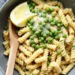 A bowl of fusilli pasta with peas, garnished with Parmesan cheese and a lemon wedge.